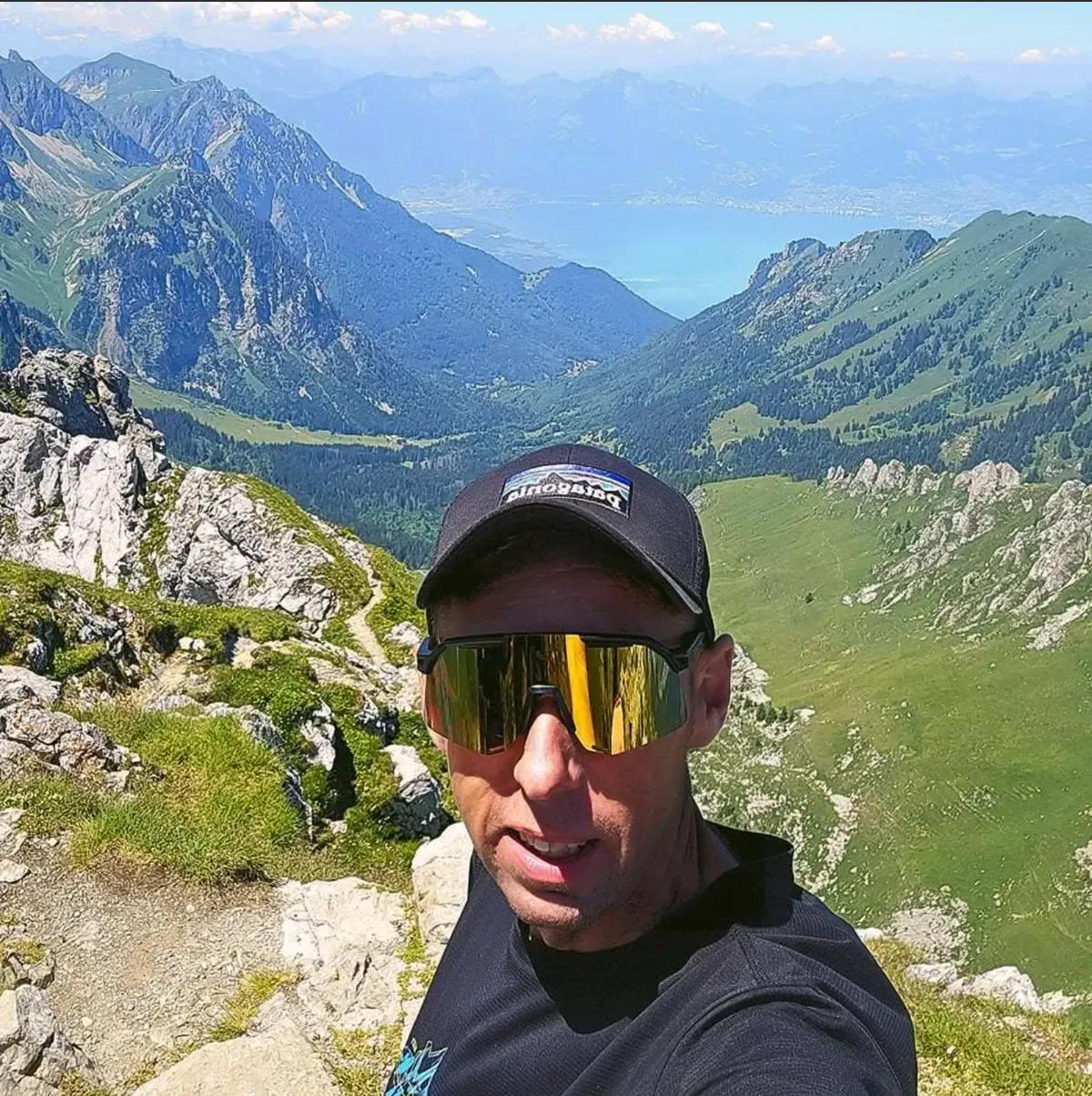 Portrait de Nicolas Descloux, fondateur d'Ô Paysage, en montagne, avec casquette et lunettes de soleil, une vallée verte en arrière-plan