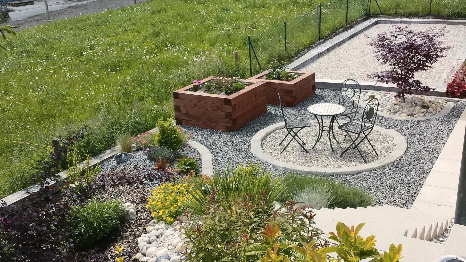 Coin repos circulaire en gravier avec table et chaises en fer forgé, bacs surélevés en bois et massifs plantés