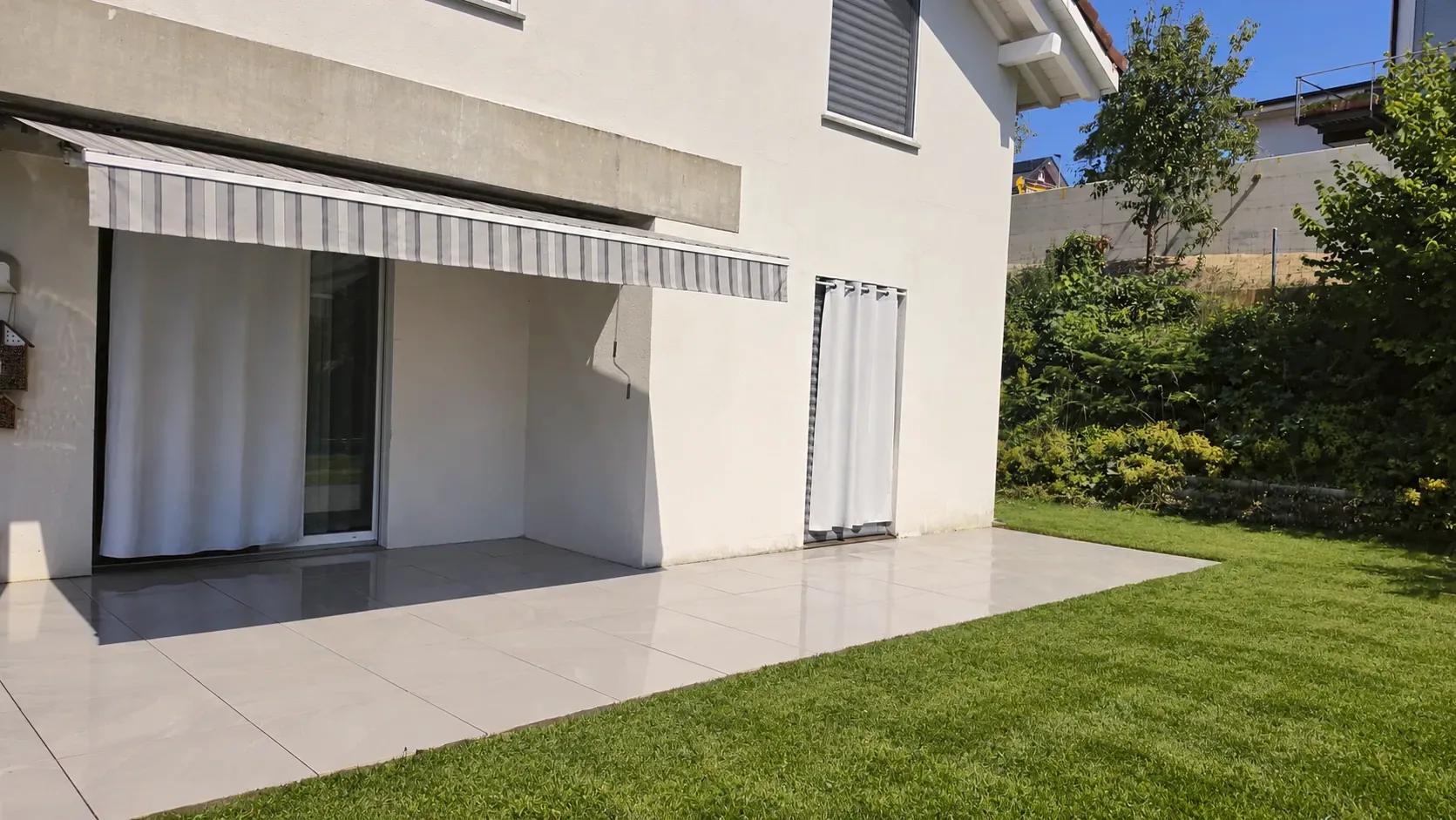 Terrasse en grès cérame posée par Ô Paysage devant une maison blanche à store banne, bordée d'une pelouse